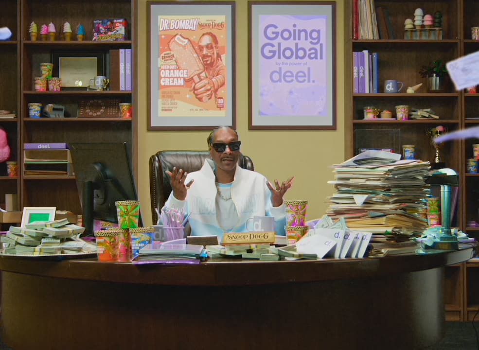 Snoop Dogg gesturing behind a desk piled with paperwork and ice cream cartons.